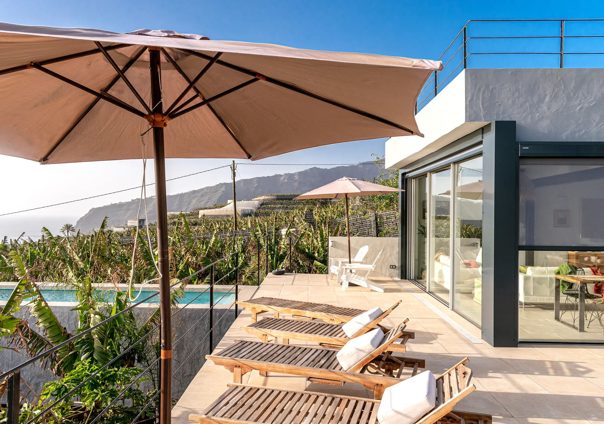 Ein moderner Außenbereich mit Liegestühlen und Sonnenschirmen auf einer Terrasse. Im Hintergrund ist ein Pool und eine Landschaft mit Hügeln und Bananenstauden. Ein Gebäude mit Glasfront ist rechts im Bild zu sehen.