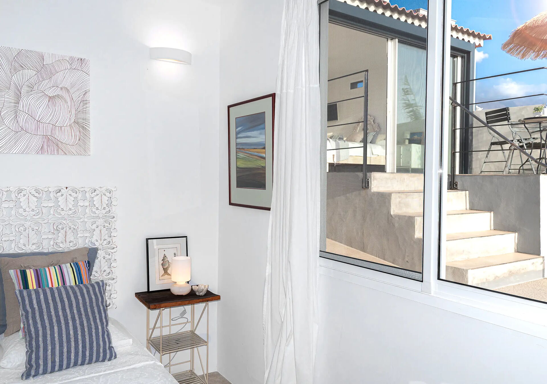 Helles Schlafzimmer mit einem weißen Bett, verzierten Kopfteil und bunten Kissen. Links steht ein kleiner Tisch mit Lampe, Kerzen, Büchern und Bildern. Rechts ein Fenster mit Blick auf eine Terrasse mit Tisch und Stühlen.