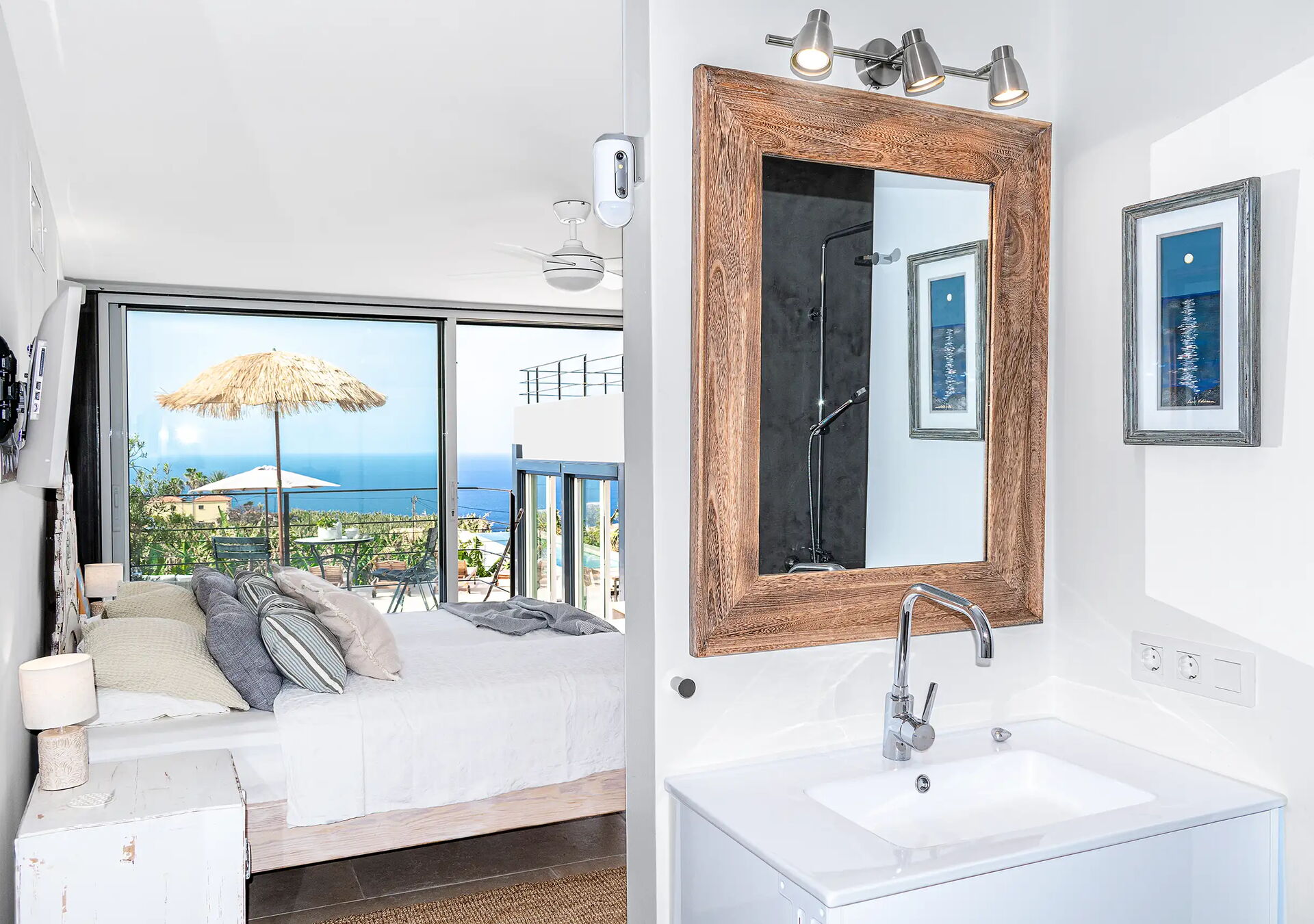 Schlafzimmer mit Meerblick zeigt ein Bett und große Fenster mit Blick auf das Meer. Links sind Kissen und ein Strohdachschirm sichtbar. Im Vordergrund ein Badezimmer mit Spiegel, Waschbecken und zwei kleinen Bildern an der Wand.
