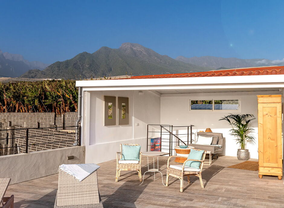 Eine überdachte Terrasse mit Sitzbereich, bestehend aus Rattanstühlen, Tisch und Sofa. Im Hintergrund sind grüne Berge sichtbar. Eine Holzkommode und eine Pflanze sind ebenfalls Teil des Arrangements. Klarer blauer Himmel.