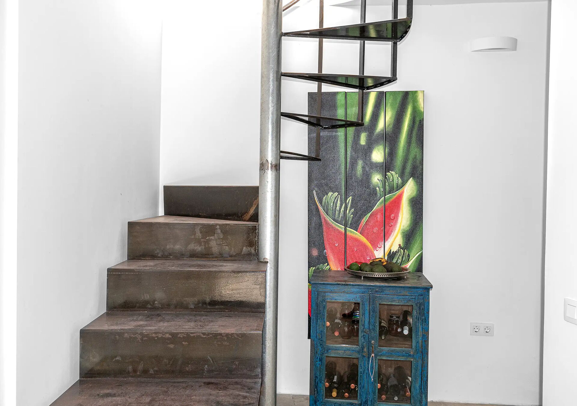 Eine kleine Wendeltreppe aus Metall steht neben einem weißen Wandsockel. Daneben befindet sich ein kleiner blauer Schrank mit Glastüren. Auf dem Schrank steht ein Gemälde mit einer roten Blume.