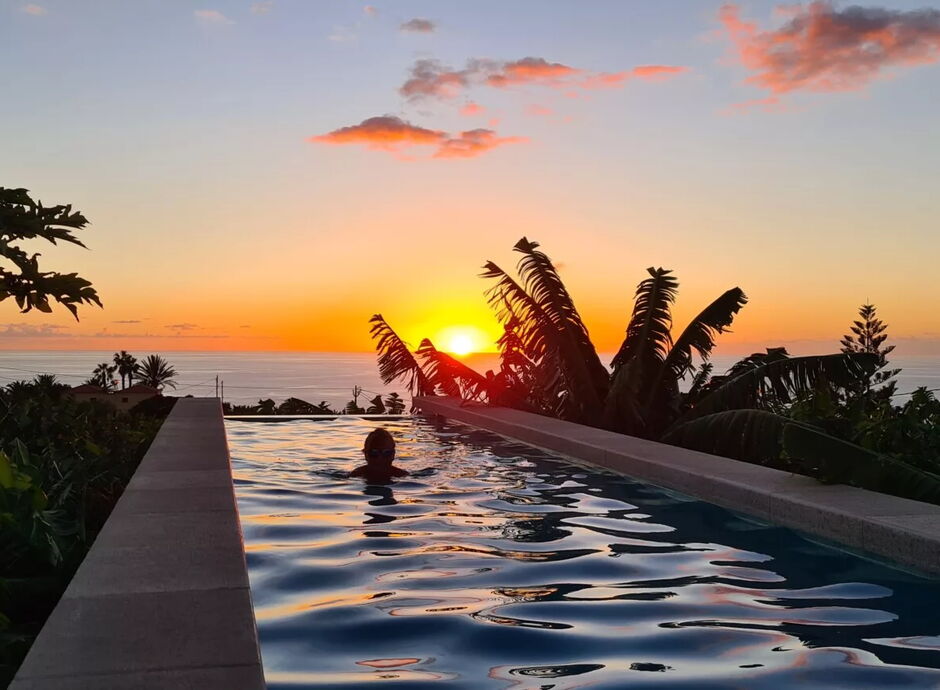 Ein Mensch schwimmt in einem Infinity-Pool, der von Pflanzen umgeben ist. Im Hintergrund ist ein Sonnenuntergang über dem Meer zu sehen, mit wenigen Wolken am Himmel. Palmen rahmen die Szenerie ein.