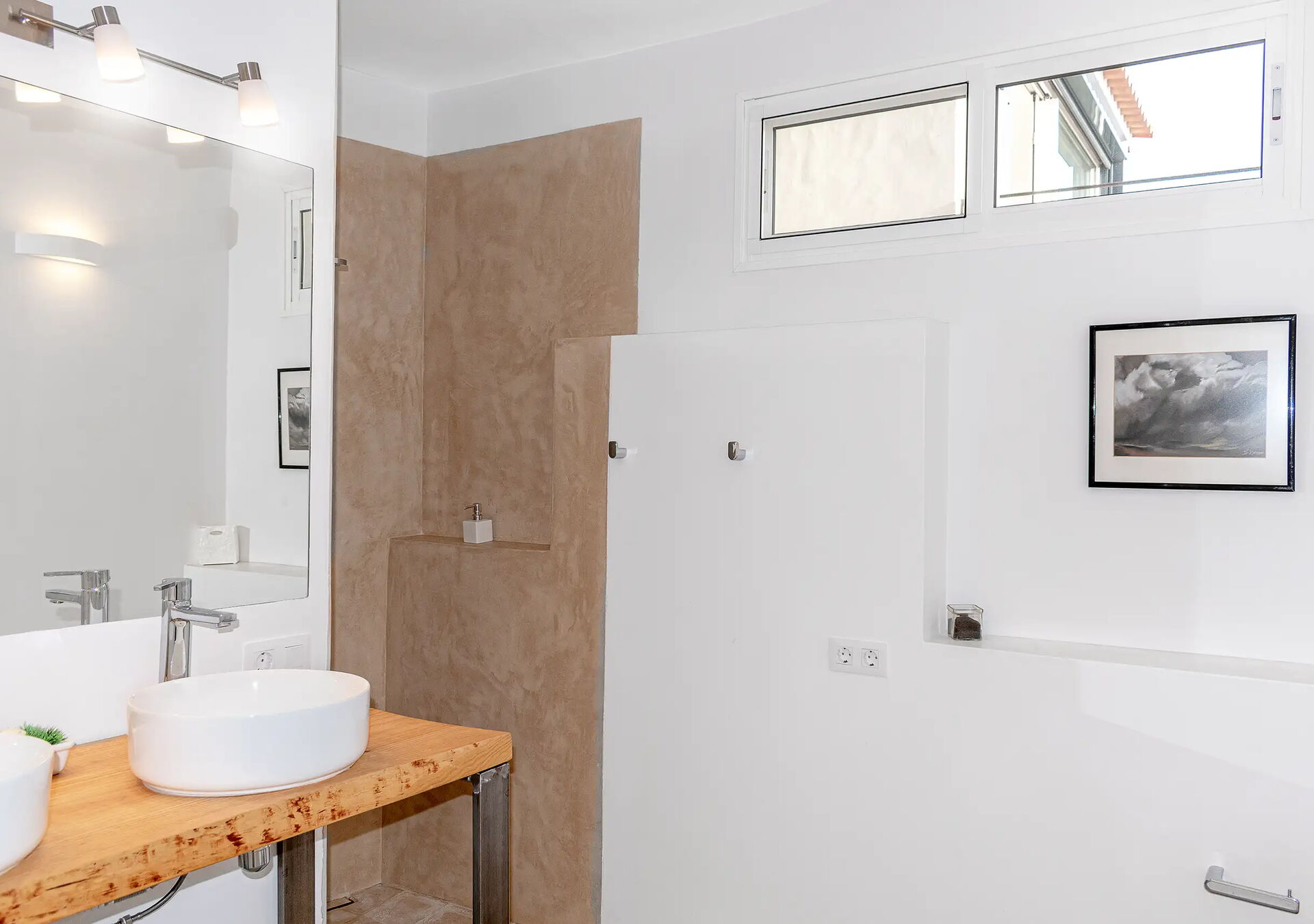 Helles Badezimmer mit weiß gestrichenen Wänden, einem großen Spiegel über einem Holztisch mit rundem Waschbecken, einer offenen Dusche mit beiger Rückwand, einem kleinen Fenster und einem gerahmten Bild an der Wand.