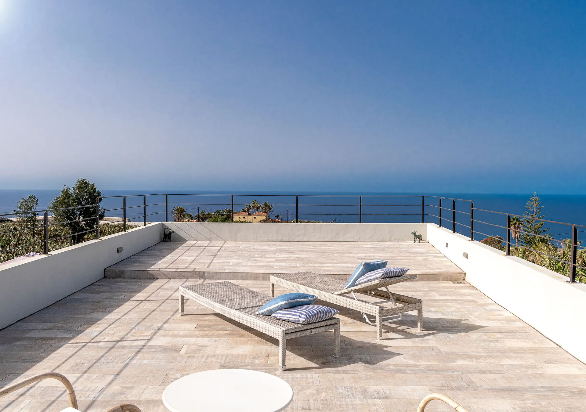 Eine erhöhte Terrasse mit zwei Liegestühlen, auf denen blaue Kissen liegen, bietet einen Blick auf das Meer und den klaren Himmel. Weiße Wände umgeben die Terrasse, und Pflanzen sind im Hintergrund zu sehen.