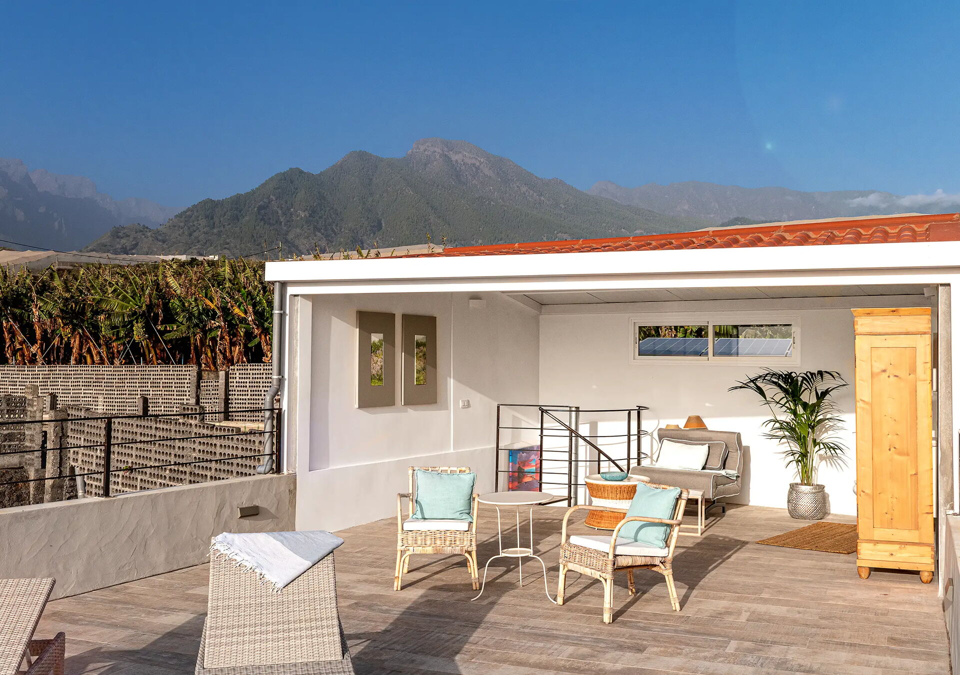 Eine überdachte Terrasse mit Sitzbereich, bestehend aus Rattanstühlen, Tisch und Sofa. Im Hintergrund sind grüne Berge sichtbar. Eine Holzkommode und eine Pflanze sind ebenfalls Teil des Arrangements. Klarer blauer Himmel.