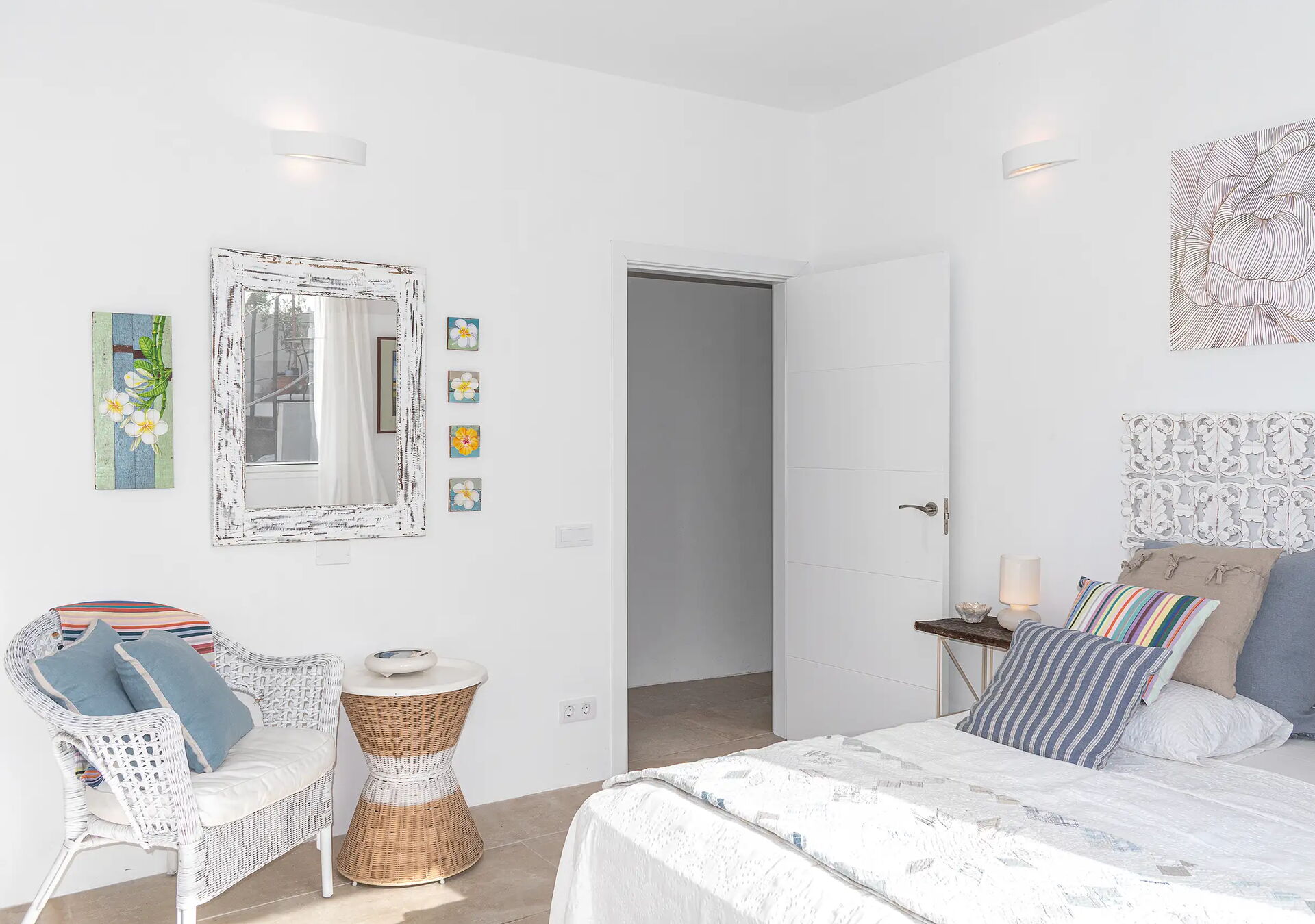 Helles Schlafzimmer mit weißem Bett, bunten Kissen und einem Nachttisch mit Lampe. Links steht ein weißer Sessel neben einem runden Beistelltisch. An den weißen Wänden hängen Kunstwerke und Fotos. Eine offene Tür führt nach draußen.