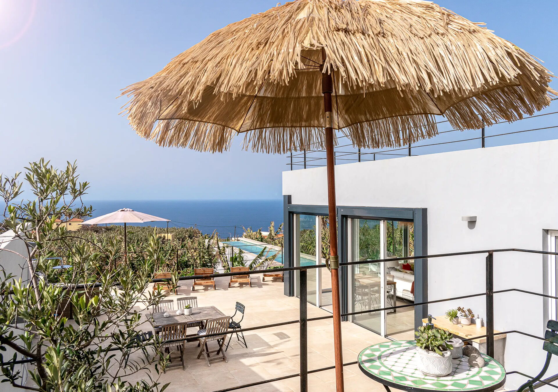 Eine Terrasse mit Strohdachschirm, Tisch und Stühlen. Moderne weiße Gebäude im Hintergrund, Meerblick in der Ferne. Pflanzen und ein Geländer aus schwarzem Metall umgeben die Fläche. Der Himmel ist klar und blau.