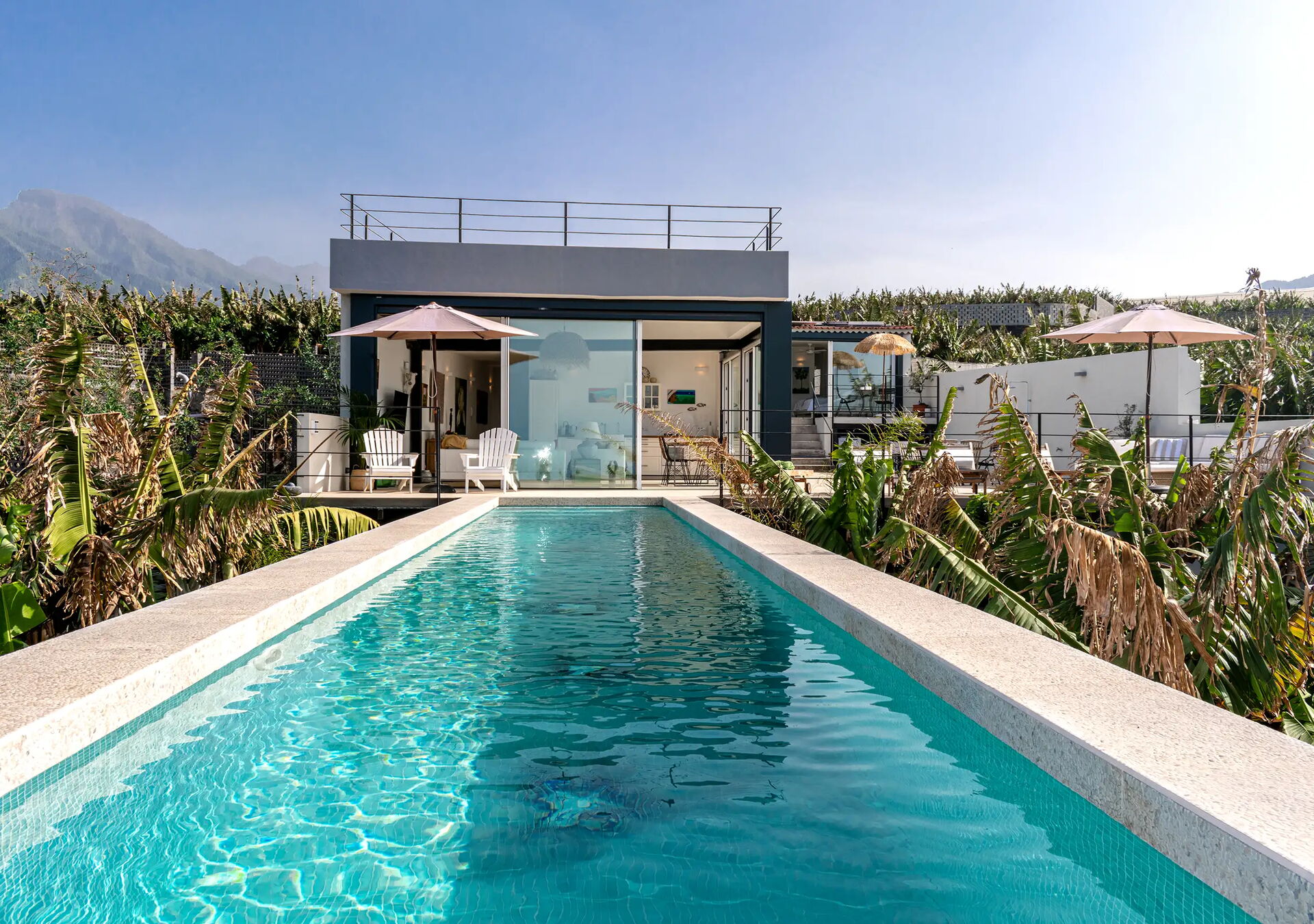 Ein modernes, flaches Gebäude mit Glasfront steht hinter einem langen Pool, der von Pflanzen umgeben ist. Im Hintergrund sind Hügel unter blauem Himmel zu sehen. Vor dem Gebäude stehen Liegestühle und Sonnenschirme.