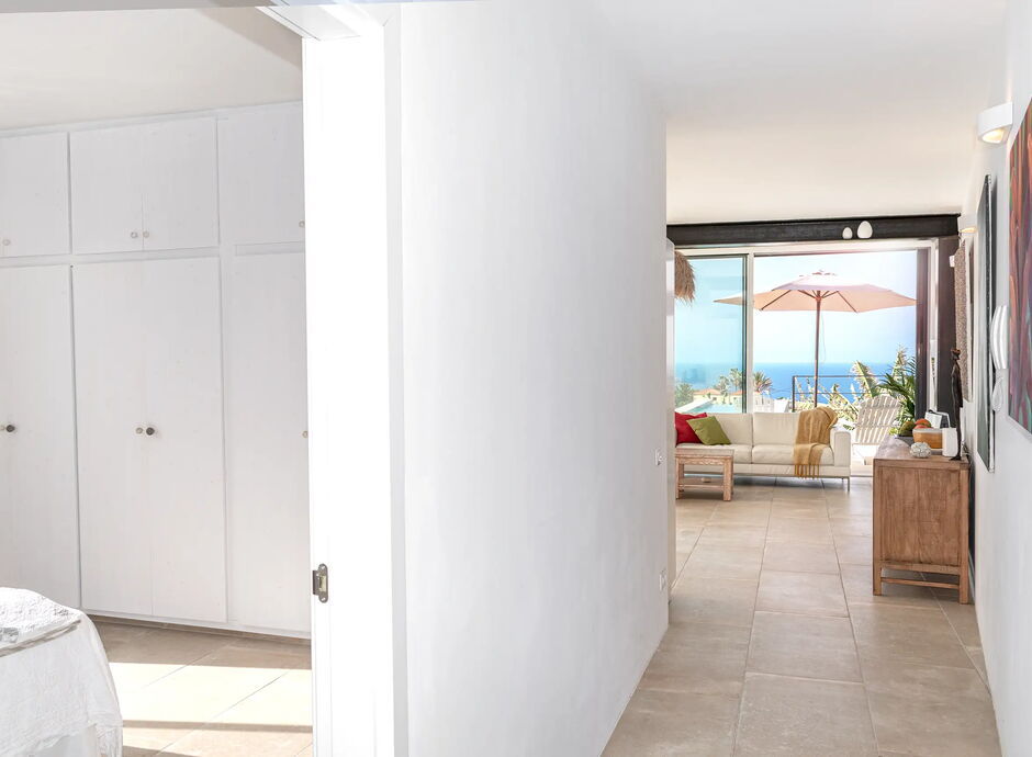 Heller Apartmentflur mit weiß gestrichenen Wänden, der zu einem Wohnzimmer führt. Große Fenster im Wohnbereich bieten Blick auf das Meer. Links sind geschlossene Kleiderschränke zu sehen. Ein Sonnenschirm ist draußen erkennbar.