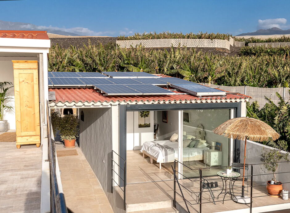 Eine moderne, weiße Ferienhütte mit großen Fenstern und Solarpaneelen auf dem Dach. Davor steht ein Tisch mit Stühlen und einem Sonnenschirm. Im Hintergrund sind viele grüne Pflanzen zu sehen.