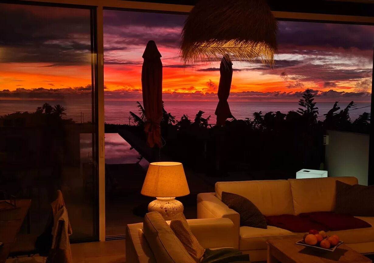 Wohnzimmer mit weißem Sofa und Lampe, hinter einer großen Glasfront. Draußen ist ein beeindruckender Sonnenuntergang zu sehen mit leuchtenden Orange-, Rot- und Lilatönen über dem Meer und silhouettierten Pflanzen.