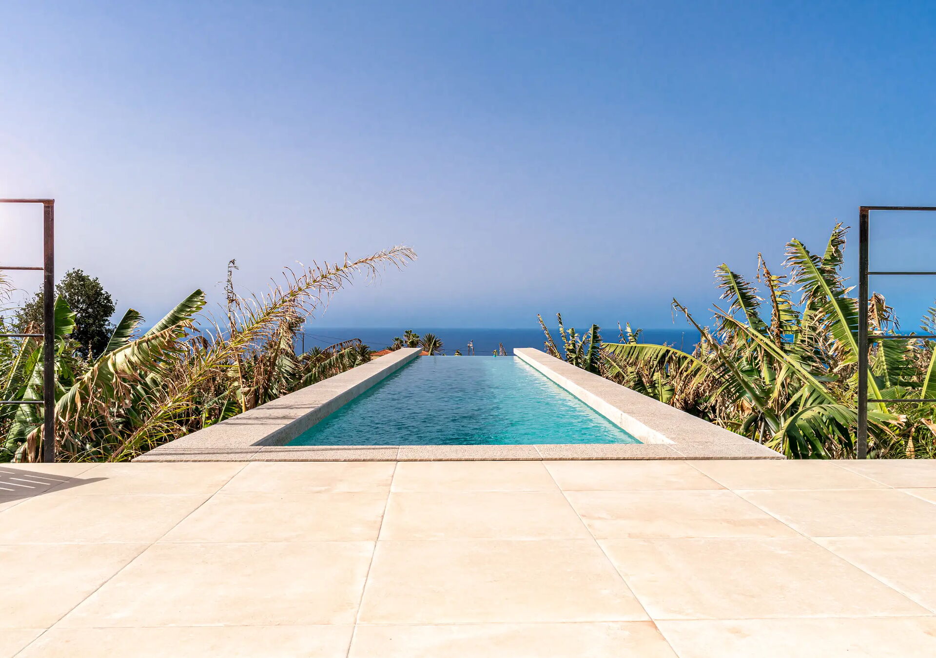 Weitläufiger Pool mit Blick auf das Meer, umgeben von grünen Pflanzen und einem klaren blauen Himmel. Der Pool erstreckt sich in Richtung Horizont und vermittelt ein Gefühl von Ruhe und Weite.