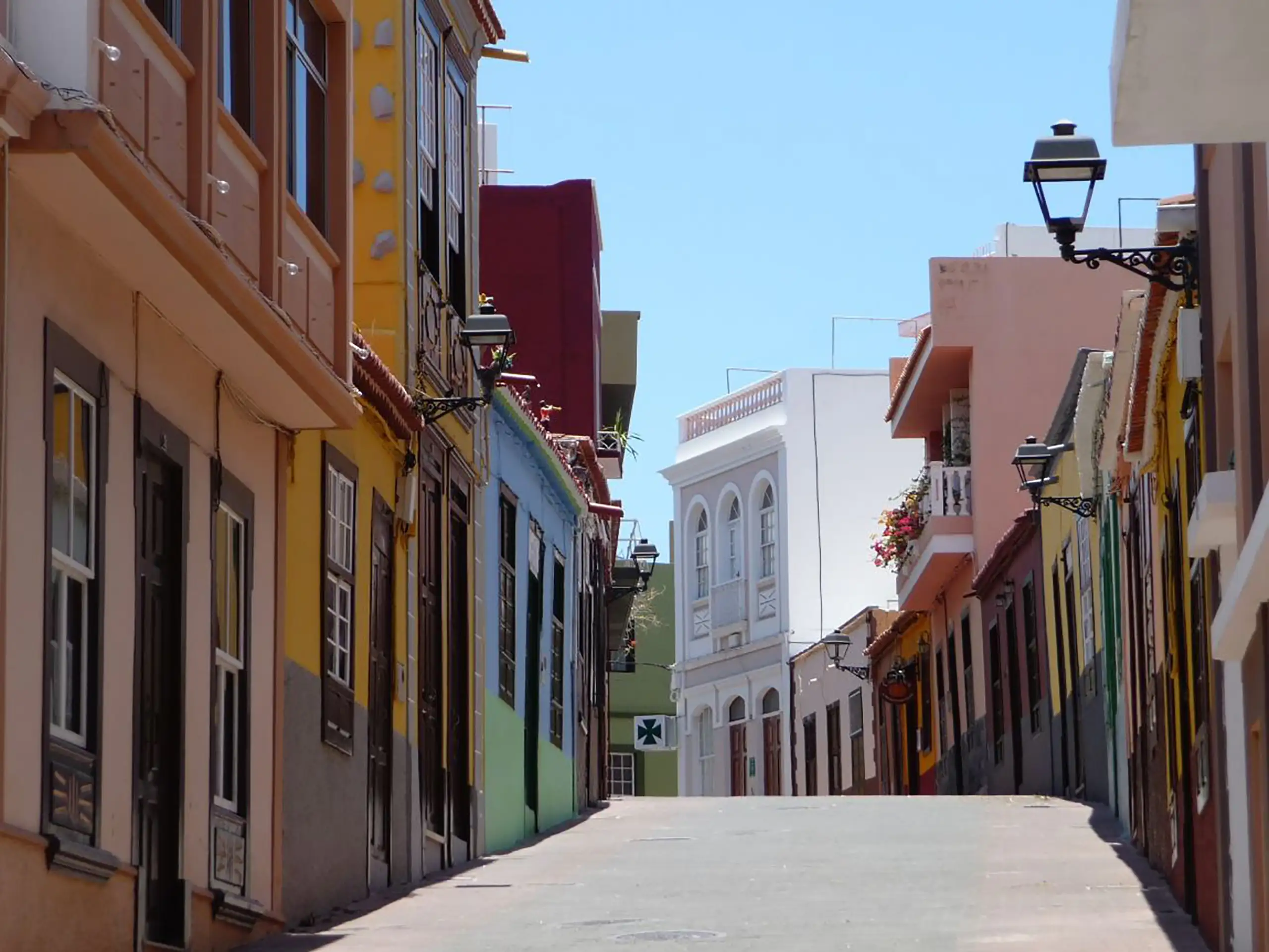 Eine enge Straße mit bunten, aneinander gereihten Häusern an beiden Seiten. Die Fassaden sind in Gelb, Rot, Blau und anderen Farben gestrichen. Laternen hängen an den Hauswänden, blauer Himmel im Hintergrund.